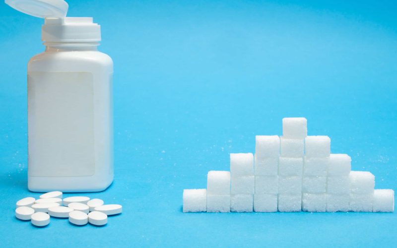 white tablets pills of sugar substitute sweetener with big bottle blank label vs pile of refined normal sugar concept on blue background