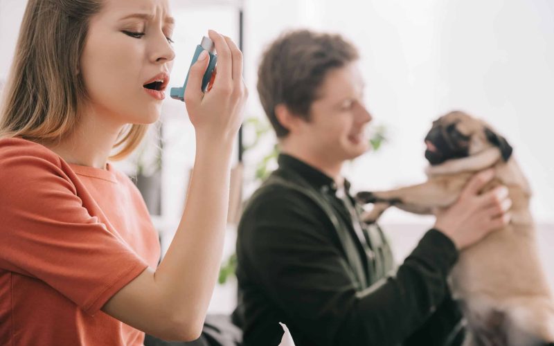 selective focus of attractive blonde woman allergic to dog using inhaler near man with pug