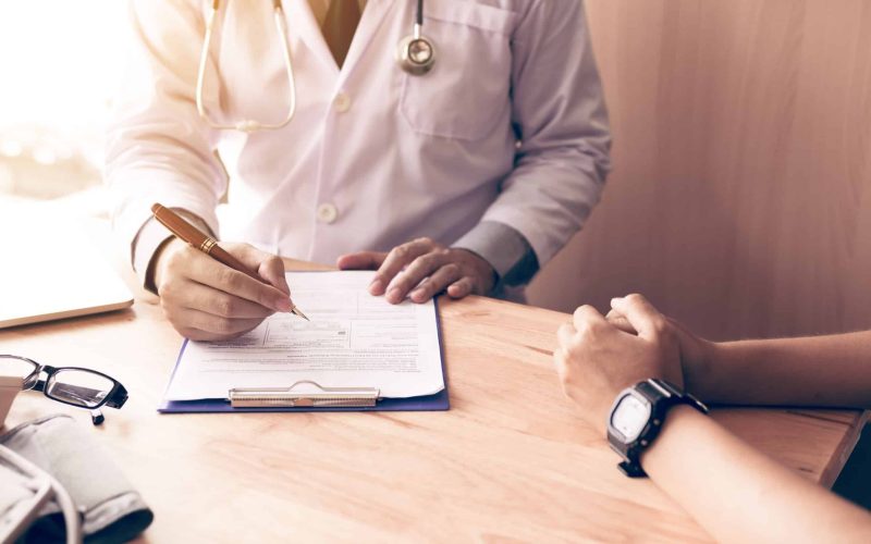 Doctor hand holding pen writing patient history list on note pad and talking to the patient about medication and treatment.