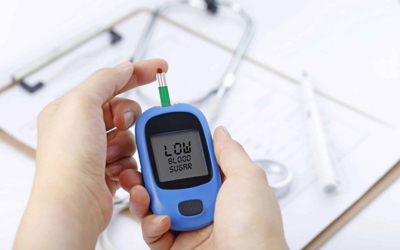 Hand holding a blood glucose meter measuring blood sugar, the background is a stethoscope and chart file